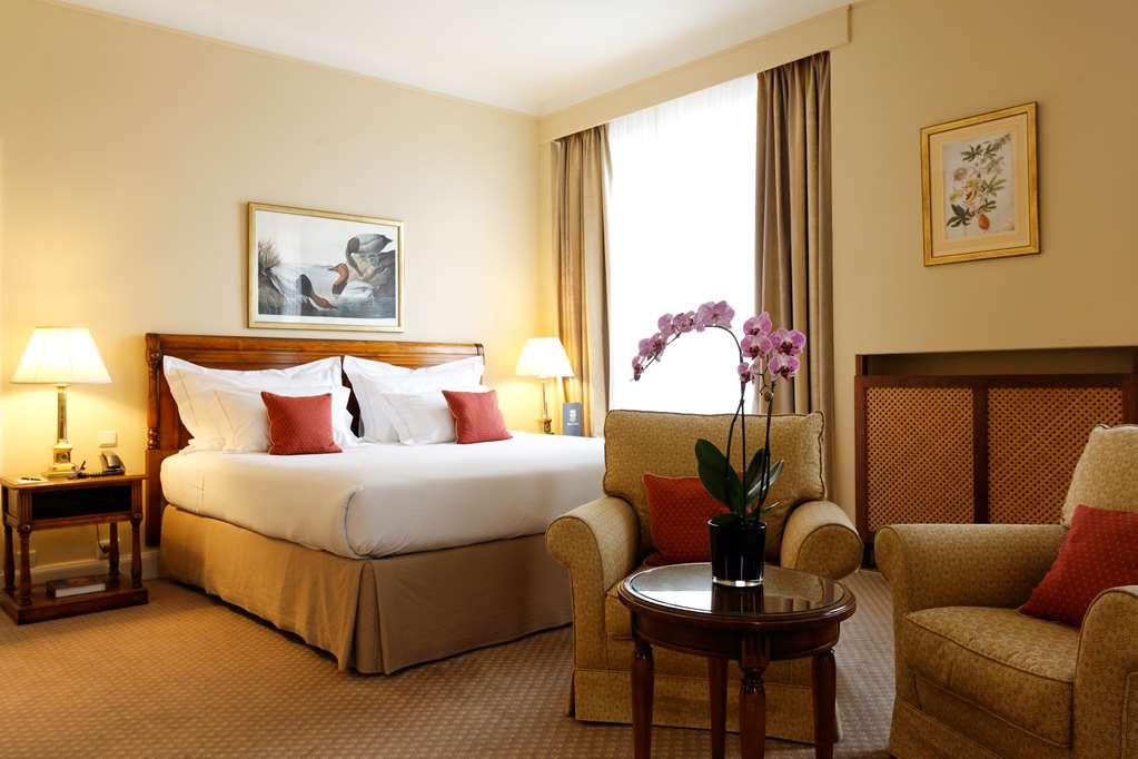 Hotel Le Plaza Brussels Exterior photo The photo shows a well-decorated hotel room. It features a large bed with white bedding and red accent pillows. There are two bedside lamps on either side of the bed. A seating area includes a comfortable armchair and a small round table with a potte
