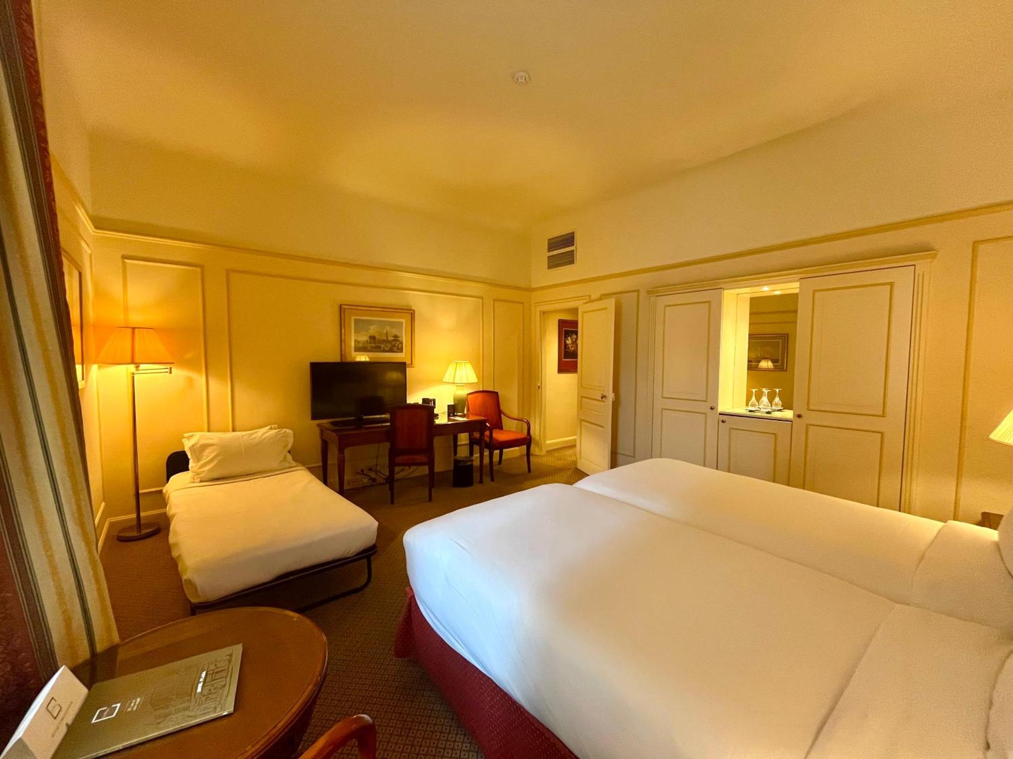 Hotel Le Plaza Brussels Room photo The photo shows an interior of a hotel room with a warm, cozy ambiance. It features two single beds made with white linens positioned side by side. There is a small, rolled-up cot or mattress in the corner. Near the beds, there is a bedside table and