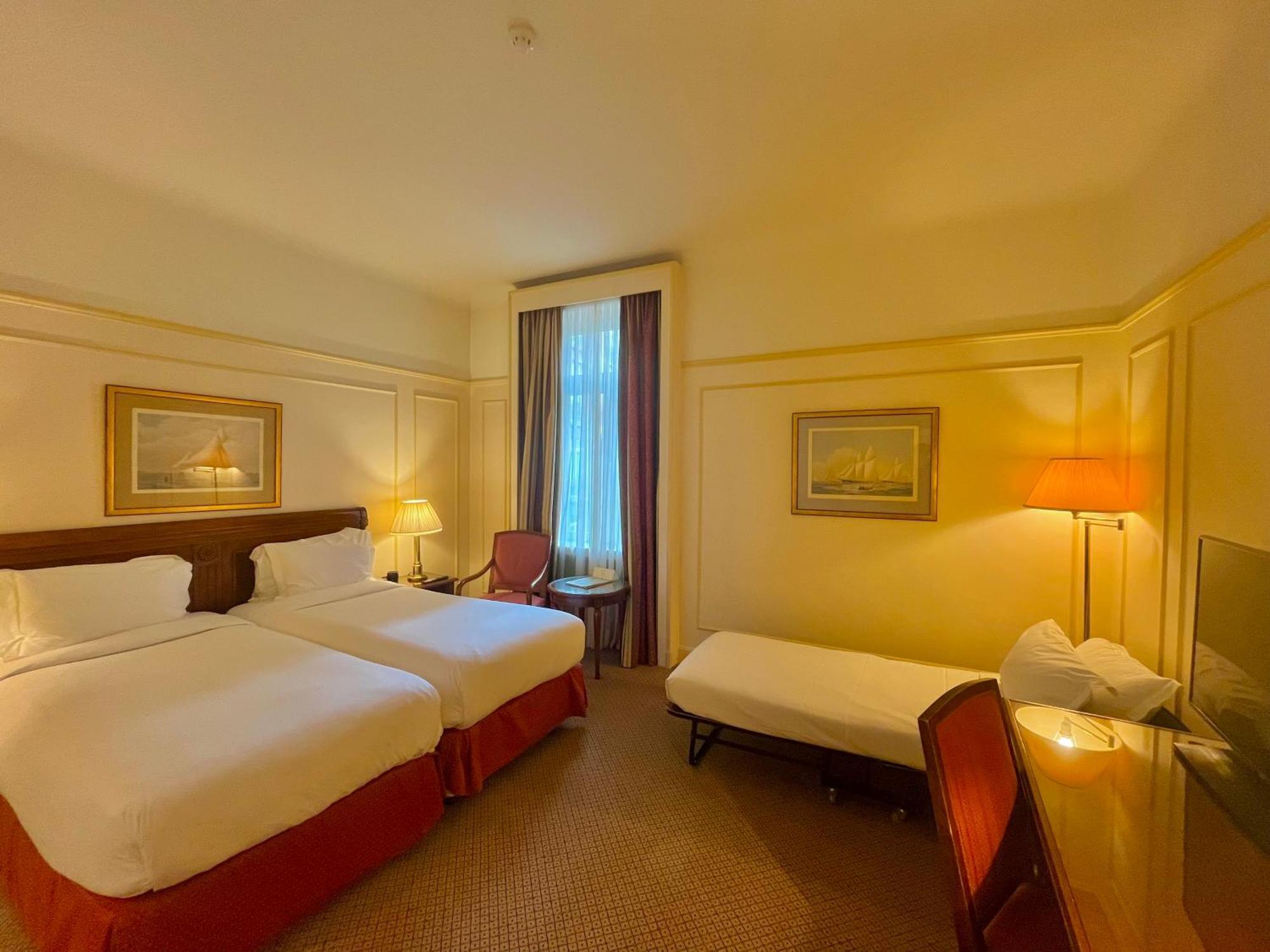 Hotel Le Plaza Brussels Room photo The photo shows a hotel room featuring two single beds with white linens and red accents. There is a small extra bed on the floor, along with a wooden chair. A desk and a lamp are present near the window, which has sheer curtains allowing natural lig